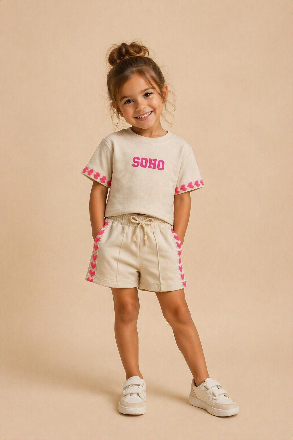 Kindermodel draagt een beige SOHO zomerset bestaande uit een comfortabel T-shirt en short met roze hartdetails langs de mouwen en broekspijpen. De outfit wordt gepresenteerd tegen een zachte beige studioachtergrond, ideaal voor meisjes die houden van trendy en comfortabele kleding.