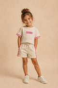 Kindermodel draagt een beige SOHO zomerset bestaande uit een comfortabel T-shirt en short met roze hartdetails langs de mouwen en broekspijpen. De outfit wordt gepresenteerd tegen een zachte beige studioachtergrond, ideaal voor meisjes die houden van trendy en comfortabele kleding.