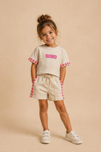 Kindermodel draagt een beige SOHO zomerset bestaande uit een comfortabel T-shirt en short met roze hartdetails langs de mouwen en broekspijpen. De outfit wordt gepresenteerd tegen een zachte beige studioachtergrond, ideaal voor meisjes die houden van trendy en comfortabele kleding.