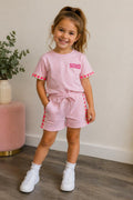 Kindmodel draagt een lichtroze Soho meisjes set bestaande uit een T-shirt en korte broek. Het T-shirt heeft de tekst "SOHO" op de borst en opvallende neonroze hartjesdetails op de mouwen. De bijpassende short heeft dezelfde hartjesdetails langs de zijkanten. De comfortabele zomerset wordt gecombineerd met witte sneakers en is gefotografeerd in een lichte, stijlvolle binnenruimte.