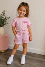 Kindmodel draagt een lichtroze Soho meisjes set bestaande uit een T-shirt en korte broek. Het T-shirt heeft de tekst "SOHO" op de borst en opvallende neonroze hartjesdetails op de mouwen. De bijpassende short heeft dezelfde hartjesdetails langs de zijkanten. De comfortabele zomerset wordt gecombineerd met witte sneakers en is gefotografeerd in een lichte, stijlvolle binnenruimte.