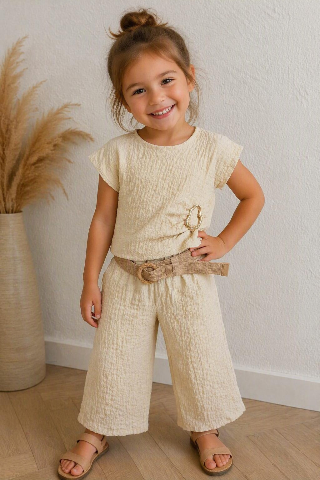 Kindmodel draagt een beige tweedelige kinder set met structuurstof, bestaande uit een korte top met decoratieve gesp, wijde broek en bijpassende riem. Comfortabele en stijlvolle zomerkleding voor kinderen van DeNa Kids Boutique.