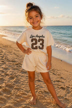 Kindmodel draagt een crème kleurig Los Angeles 23 kinder setje met luipaardprint op het strand. De tweedelige zomerset bestaat uit een comfortabele oversized T-shirt en bijpassende short uit de collectie van DeNa Kids Boutique.