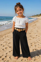 Kindermodel draagt een stijlvolle zwart-witte kinder jumpsuit met witte off-shoulder top, lange zwarte broek en rieten riem op een zonnig strand. Comfortabele en trendy zomerkleding voor kinderen.