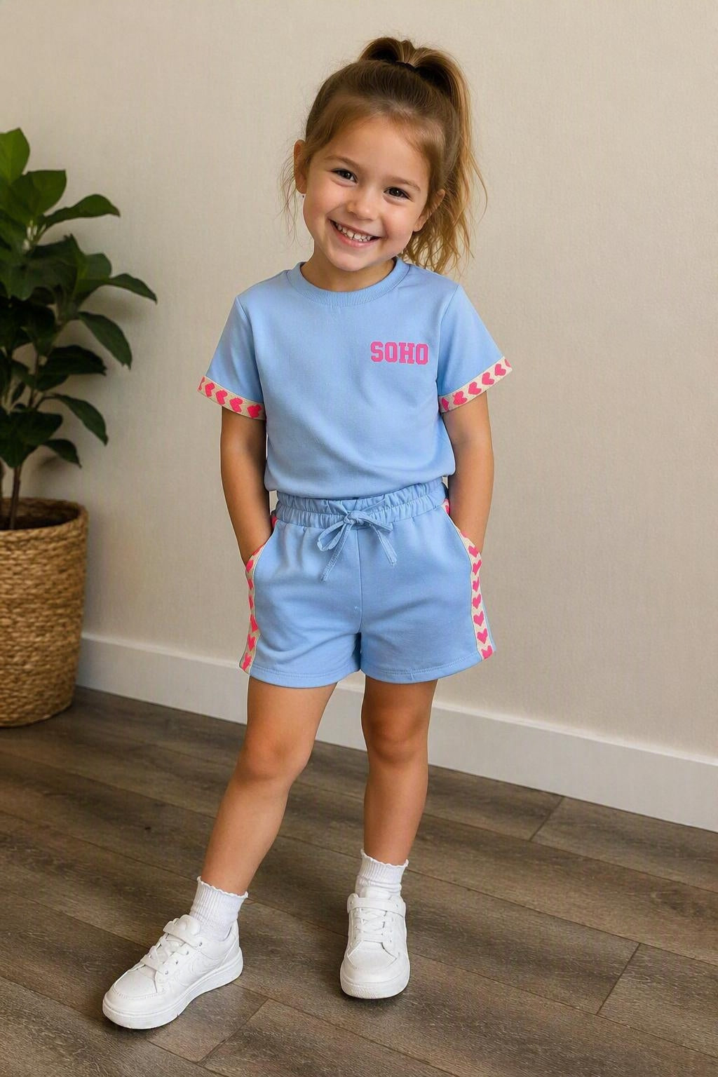 Kindmodel draagt een lichtblauw SOHO zomersetje met T-shirt en korte broek, afgewerkt met roze hartjesdetails op de mouwen en zijkanten, gecombineerd met witte sneakers in een neutrale webshopsetting.