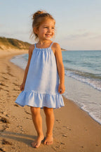 Kindermodel draagt een lichtblauw linnen kinderjurkje met verstelbare schouderbandjes en volantzoom op het strand tijdens een zonnige zomerdag. Perfect zomerjurkje voor kinderen, comfortabel, luchtig en stijlvol voor vakantie, strand en warme dagen.
