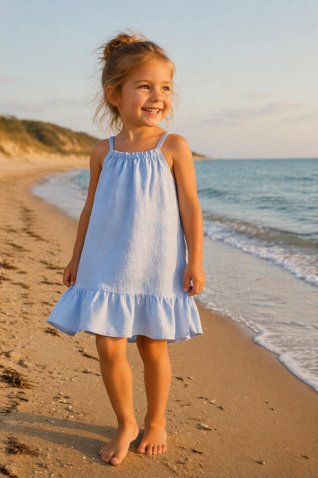 Kindermodel draagt een lichtblauw linnen kinderjurkje met verstelbare schouderbandjes en volantzoom op het strand tijdens een zonnige zomerdag. Perfect zomerjurkje voor kinderen, comfortabel, luchtig en stijlvol voor vakantie, strand en warme dagen.