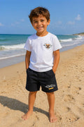 Kindermodel draagt een wit Diamond Club T-shirt met zwarte korte broek op het strand. Comfortabel en stoer kinder zomersetje in wit en zwart voor jongens, verkrijgbaar in maten 4 tot en met 14.