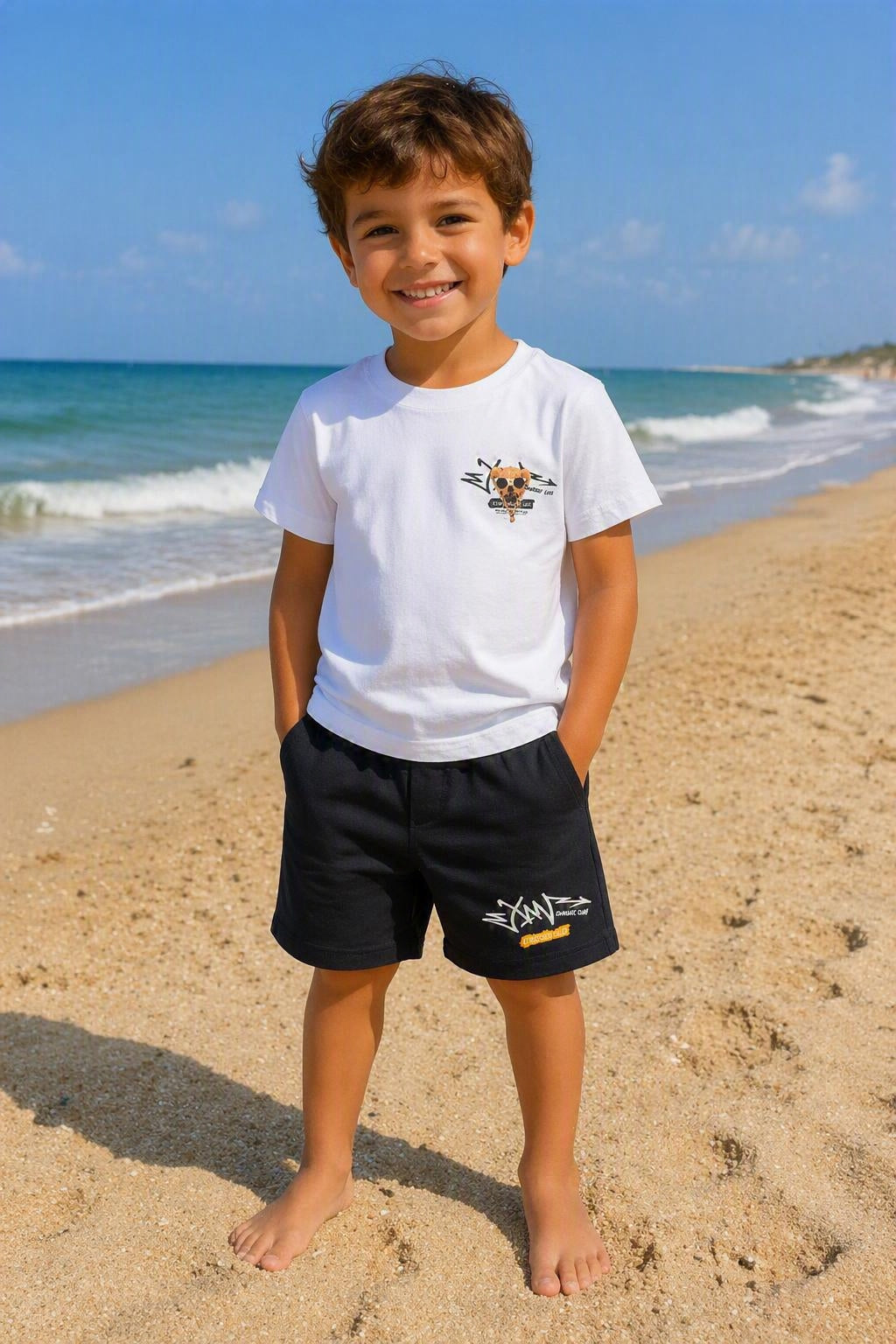 Kindermodel draagt een wit Diamond Club T-shirt met zwarte korte broek op het strand. Comfortabel en stoer kinder zomersetje in wit en zwart voor jongens, verkrijgbaar in maten 4 tot en met 14.