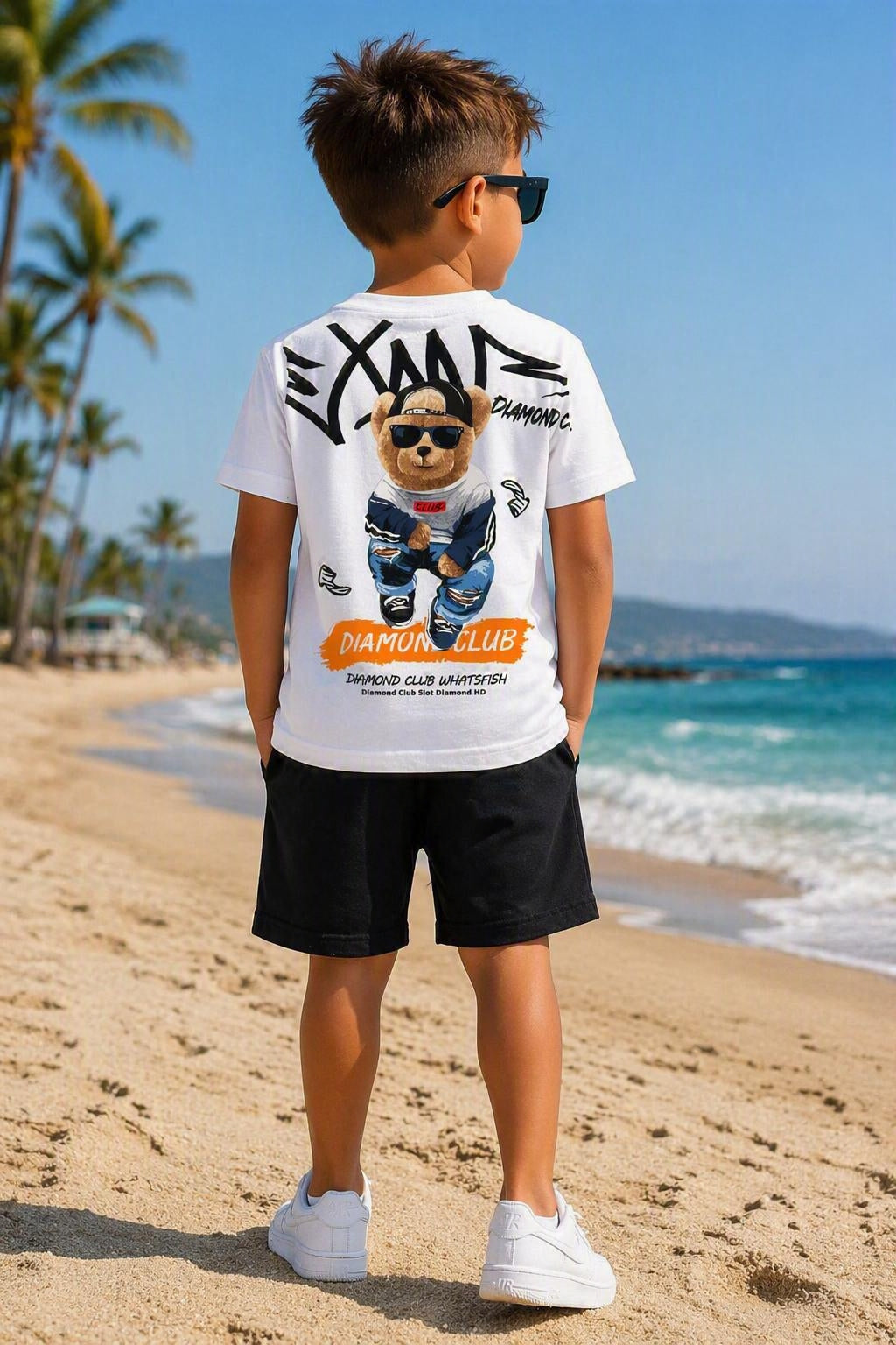 Kindermodel op het strand met de achterkant van een wit Diamond Club T-shirt met grote teddybeerprint op de rug en een zwarte korte broek, zomers jongens setje uit de collectie van DeNa Kids Boutique.