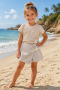 Beige kinderoutfit met een ribstof T-shirt en overslagrokje-short met decoratieve bloemen, gedragen door een kindmodel op een zonnig zandstrand aan zee. De luchtige zomerset heeft een stijlvolle neutrale kleur en een comfortabele pasvorm, ideaal voor warme dagen en vakanties.