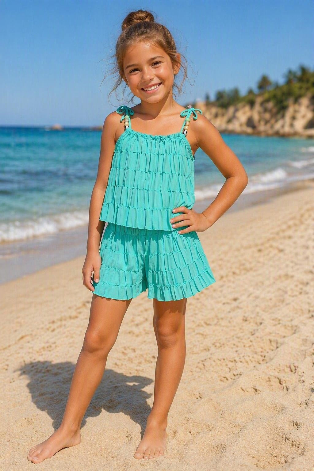 Turquoise zomerset voor kinderen bestaande uit een luchtige top met verstelbare schouderbandjes en bijpassende korte broek met ruches. De set wordt gedragen door een kindmodel op een zonnig strand met zee en zand op de achtergrond. Perfect voor warme zomerdagen, vakanties en stranduitjes.