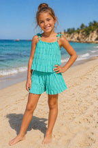 Turquoise zomerset voor kinderen bestaande uit een luchtige top met verstelbare schouderbandjes en bijpassende korte broek met ruches. De set wordt gedragen door een kindmodel op een zonnig strand met zee en zand op de achtergrond. Perfect voor warme zomerdagen, vakanties en stranduitjes.