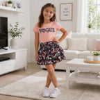 Kindermodel draagt een roze meisjes set met een T-shirt voorzien van de tekst "VOGUE" en een donkerblauw bloemenrokje met roze, witte en rode bloemen. De foto is gemaakt in een lichte woonkamer met witte accenten, waaronder een witte bank, salontafel en decoratieve accessoires. Perfecte webshopfoto voor meisjesmode en zomerkleding.
