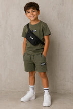 Groen ICON kinderset bestaande uit een T-shirt en korte broek met zwarte ICON-details, gedragen door een kindmodel met bijpassende zwarte crossbody tas en witte sneakers. Comfortabele en stijlvolle zomeroutfit voor kinderen, geschikt voor dagelijks gebruik, school en vrije tijd. Verkrijgbaar in de maten 4, 6, 8, 10, 12 en 14.