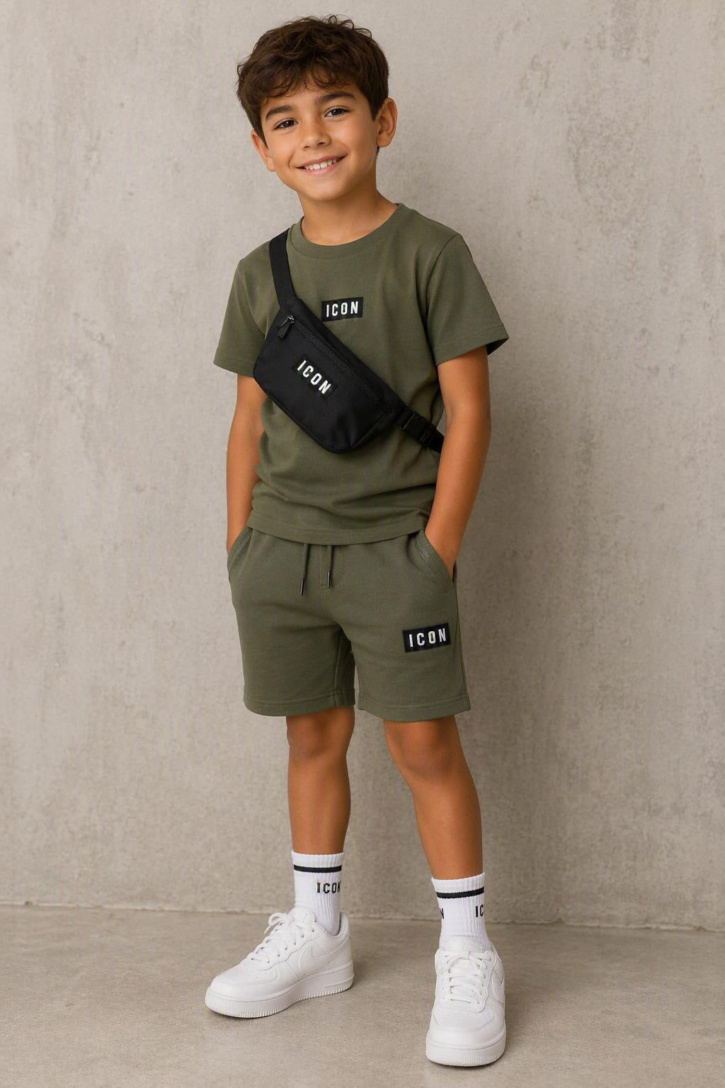 Groen ICON kinderset bestaande uit een T-shirt en korte broek met zwarte ICON-details, gedragen door een kindmodel met bijpassende zwarte crossbody tas en witte sneakers. Comfortabele en stijlvolle zomeroutfit voor kinderen, geschikt voor dagelijks gebruik, school en vrije tijd. Verkrijgbaar in de maten 4, 6, 8, 10, 12 en 14.