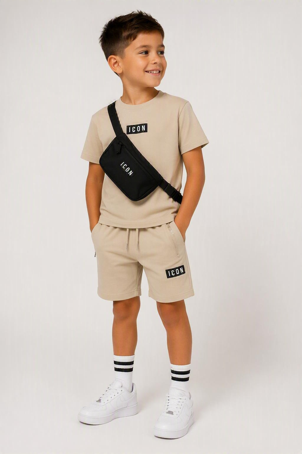 Beige ICON kinderset bestaande uit een T-shirt en korte broek met zwarte ICON-details, gedragen door een kindmodel met bijpassende zwarte crossbody tas en witte sneakers. Comfortabele en stijlvolle zomeroutfit voor kinderen, geschikt voor dagelijks gebruik, school en vrije tijd. Verkrijgbaar in de maten 4, 6, 8, 10, 12 en 14.