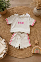 Beige meisjes set bestaande uit een oversized korte mouwen sweater met roze SOHO-opdruk en bijpassende short met roze hartjesdetails. De outfit ligt stijlvol gepresenteerd op een natuurlijke beige jute ondergrond met decoratieve accessoires in warme neutrale tinten, perfect voor een luxe kinderkleding webshop.