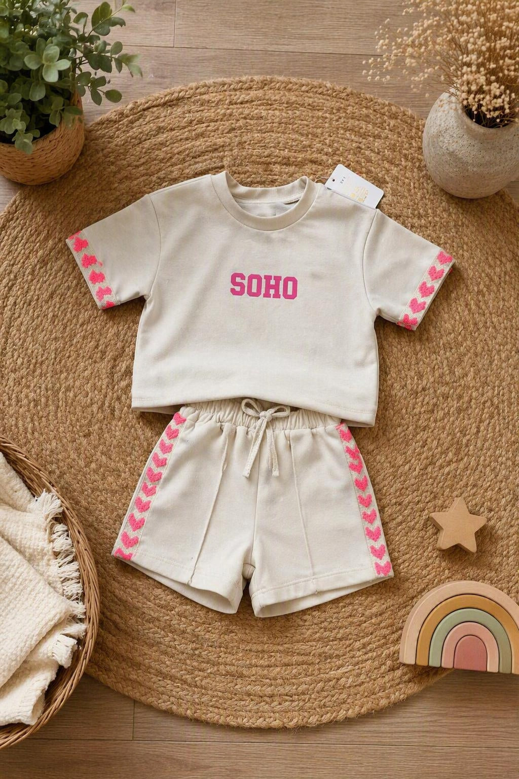 Beige meisjes set bestaande uit een oversized korte mouwen sweater met roze SOHO-opdruk en bijpassende short met roze hartjesdetails. De outfit ligt stijlvol gepresenteerd op een natuurlijke beige jute ondergrond met decoratieve accessoires in warme neutrale tinten, perfect voor een luxe kinderkleding webshop.