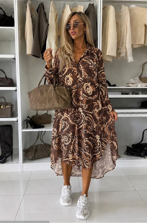 Bruine midi jurk met paisleyprint, lange mouwen en soepelvallende pasvorm gecombineerd met witte sneakers en een bruine handtas.