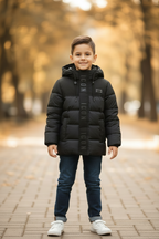Jongen in zwarte ICON gewatteerde winterjas met capuchon en donkerblauwe jeans, lifestyle bokeh achtergrond - DeNa Fashion